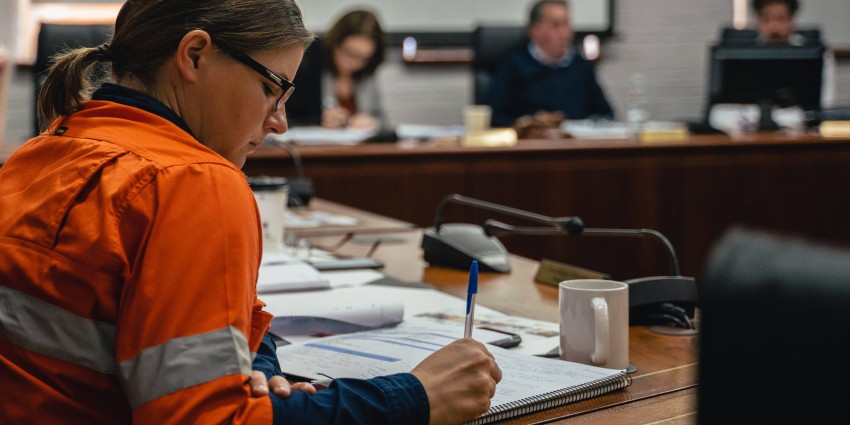 women with glasses in orange uniform writing at a table. Three people sit in the background out of focus.