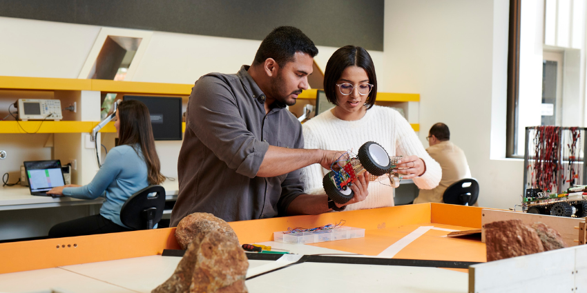 two people discussing a mechanical robotics part in a classroom environment with rocks on the table in the foreground and people working on computers in the background