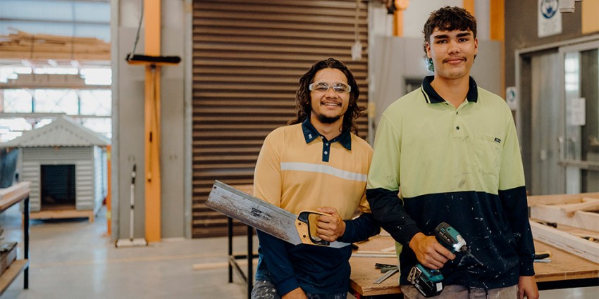 Two young male apprentices in work attire, smiling in a workshop.
