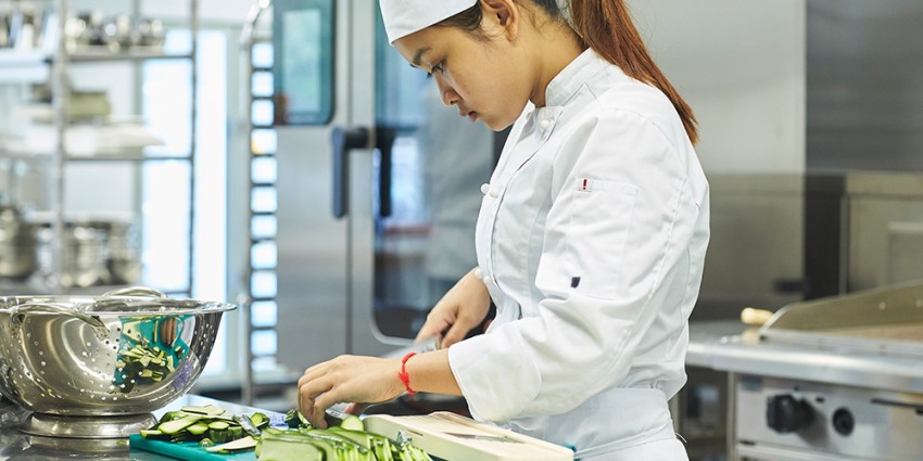 A young female student chef in a white uniform and hat preparing vegetables in a commercial kitchen. 