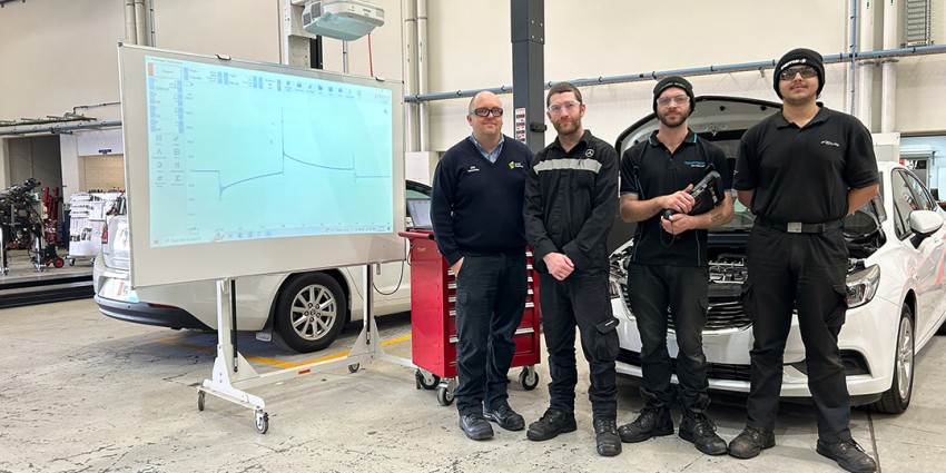 Group of four mechanics standing in an automotive workshop beside a car and a projected diagnostic chart.