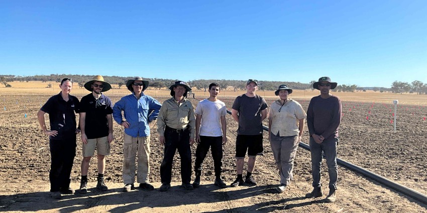 Eight people standing in a row in a dry paddock.