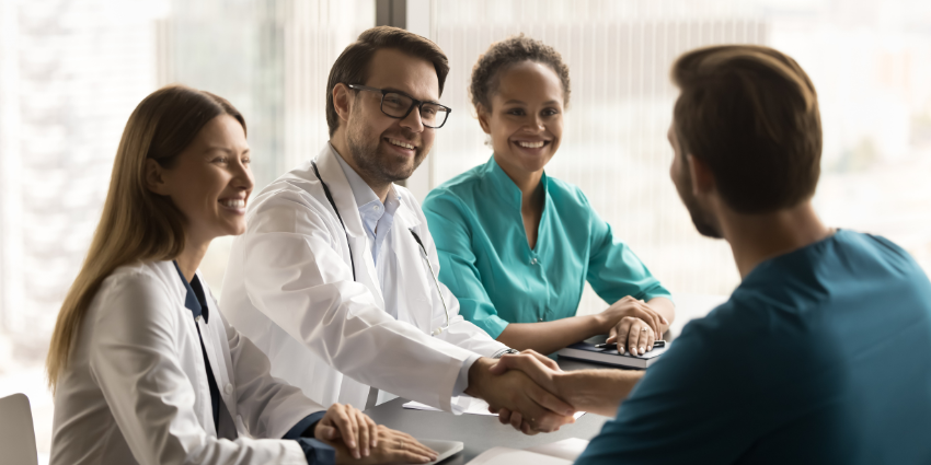 Image of two women and two men seated at a table. One of the men, dressed in a white medical coat, is shaking hands with the other man.