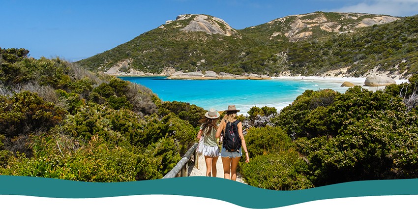 Two girls walking on a boardwalk to a secluded beach surrounded by green bush/hills