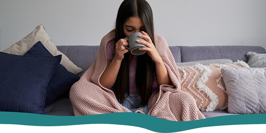 A person wrapped in a blanket and drinking from a cup on the sofa