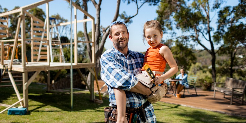 A man in a plaid shirt and tool belt holds a young girl in an orange top, with a construction project in progress behind them in a sunny outdoor setting.