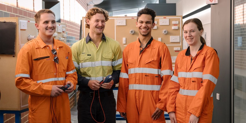 Four students in safety uniforms smiling in a training facility with electrical equipment in the background.