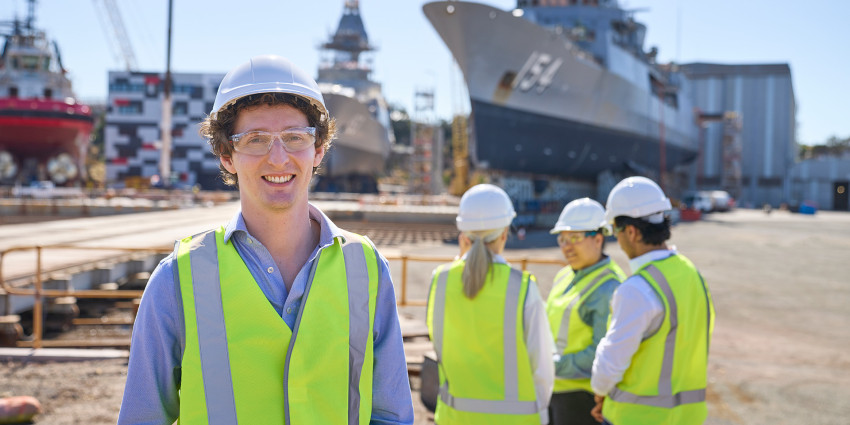 Person in a high-visibility vest and hard hat smiling in a shipyard, with colleagues and large ships in the background.