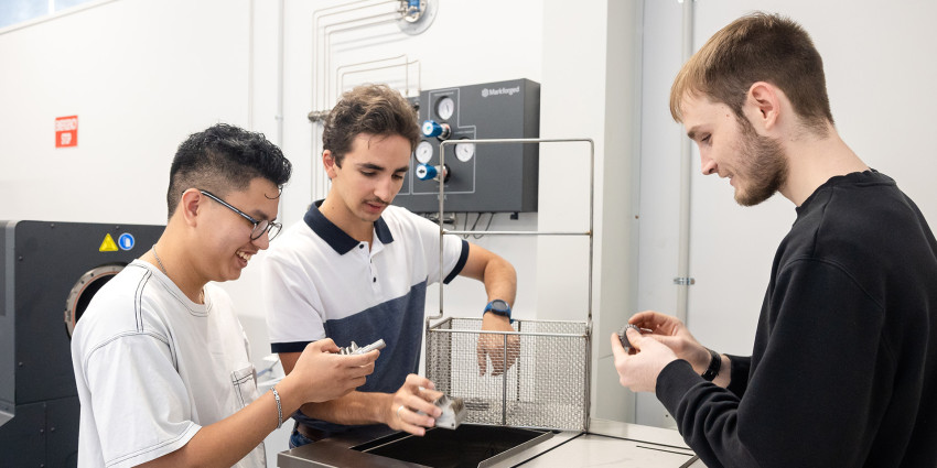 Three young individuals examining 3D-printed components near a Markforged Wash-1 machine in a technical lab setting.