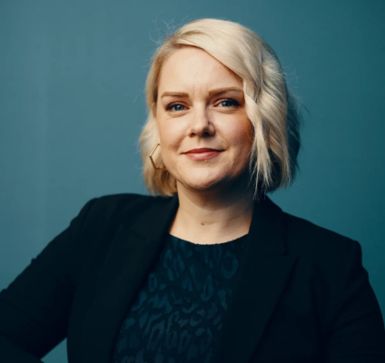 middle aged white woman with bright blonde hair in a bob cut, curled wearing black blazer and smiling against teale background