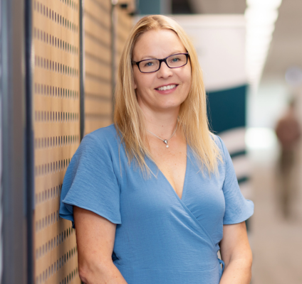 blonde woman with dark glasses wearing blue dress and standing in a corridor smiling at camera