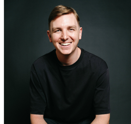 white man with light brown hair smiling in a black tshirt against a dark background